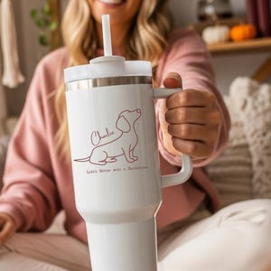 May include: White insulated tumbler with a handle and a straw. The tumbler features a brown outline of a dachshund dog, the name "Charlie", and the text "Life's better with a Dachshund."