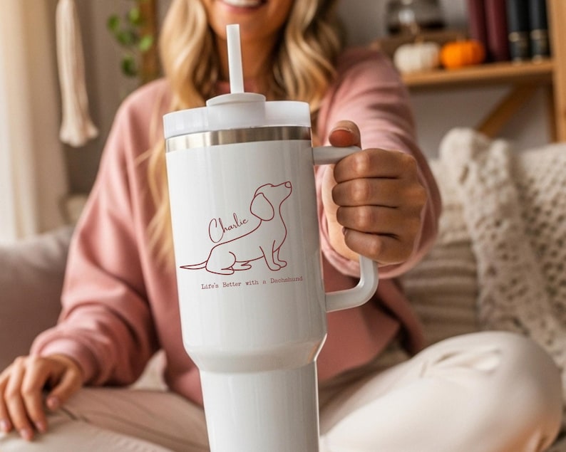 May include: White insulated tumbler with a handle and a straw. The tumbler features a brown outline of a dachshund dog, the name "Charlie", and the text "Life's better with a Dachshund."