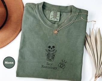 May include: A moss green t-shirt with a crew neck and short sleeves. The shirt features a black graphic of a skull and crossbones with the words "Say: Radiology" and a camera icon. A gold necklace rests on the shirt.