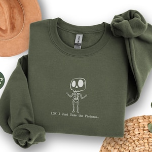 May include: Military green sweatshirt with a white skeleton graphic and the text "IDK I Just Take the Pictures...". A brown hat is visible in the upper left corner. The sweatshirt is folded with one sleeve bunched up.