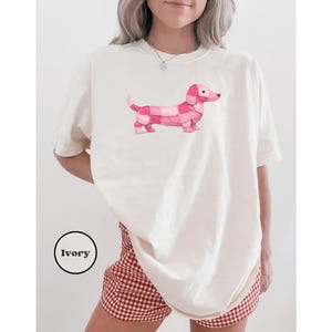 May include: Ivory-colored t-shirt featuring a pink dachshund design. The dachshund is made up of various shades of pink squares. The shirt is a relaxed fit. The word "Ivory" is in a black circle in the lower left corner.