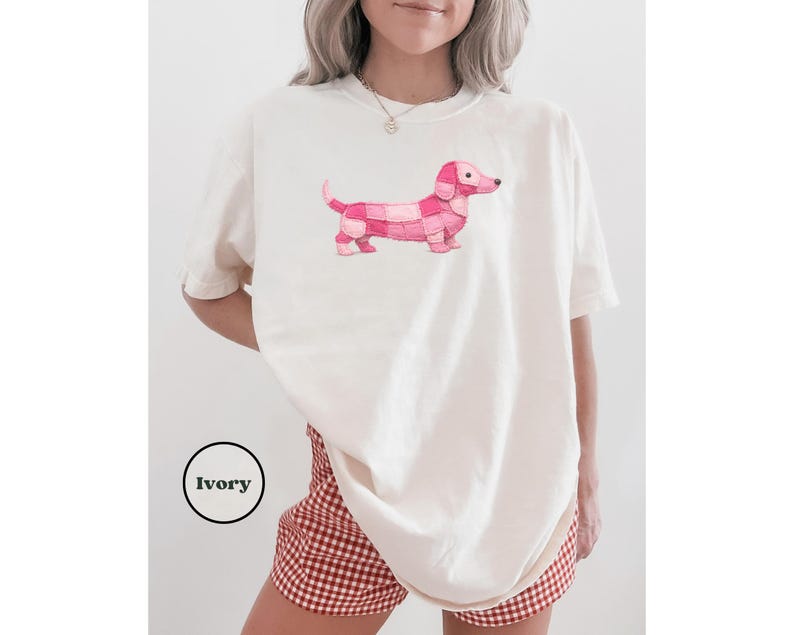 May include: Ivory-colored t-shirt featuring a pink dachshund design. The dachshund is made up of various shades of pink squares. The shirt is a relaxed fit. The word "Ivory" is in a black circle in the lower left corner.