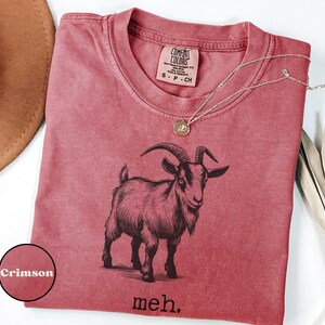 May include: A crimson red t-shirt with a black and white illustration of a goat and the text "meh."