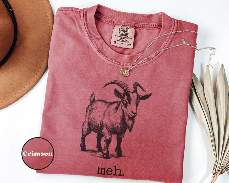 May include: A crimson red t-shirt with a black and white illustration of a goat and the text "meh."