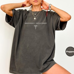 May include: A charcoal grey oversized t-shirt featuring the text EMERGENCY DEPARTMENT in white capital letters above a white caduceus symbol. The shirt has a crew neckline, short sleeves, and a relaxed fit. A small circular label reads Pepper.