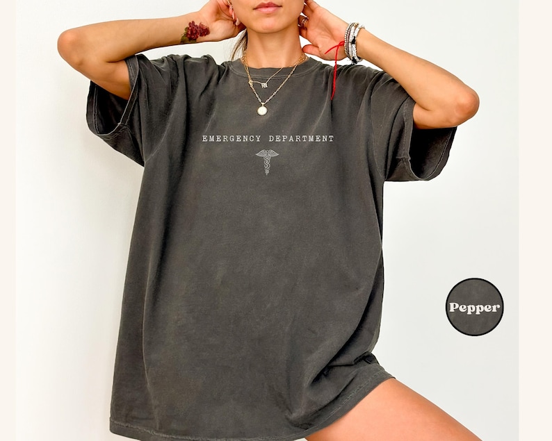 May include: A charcoal grey oversized t-shirt featuring the text EMERGENCY DEPARTMENT in white capital letters above a white caduceus symbol. The shirt has a crew neckline, short sleeves, and a relaxed fit. A small circular label reads Pepper.
