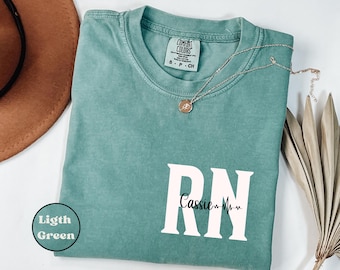 May include: A light green t-shirt with the text "RN" in white, with the name "Cassie" and a heartbeat graphic underneath. The shirt is folded and laid flat on a white surface.