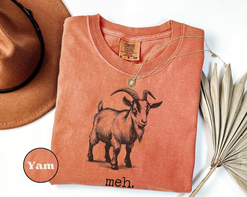 May include: A burnt orange t-shirt with a black and white illustration of a goat. The shirt has the text "yam" and "meh." printed on it.