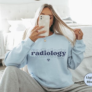 May include: Light blue sweatshirt with the word "radiology" in navy blue script across the chest. Below the word is a small navy blue radiation symbol. The sweatshirt is a crew neck style.
