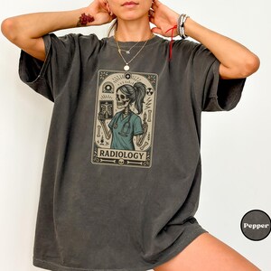 May include: Dark gray t-shirt featuring a graphic of a skeleton radiologist holding an X-ray. The graphic is framed with decorative elements and the word "RADIOLOGY" at the bottom. The shirt has short sleeves and a relaxed fit.