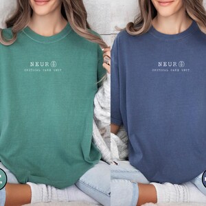 May include: Two short-sleeved t-shirts, one light green and one denim blue. Each shirt has the word "NEUR" above "CRITICAL CARE UNIT" in white text. The shirts are worn with light-wash jeans.