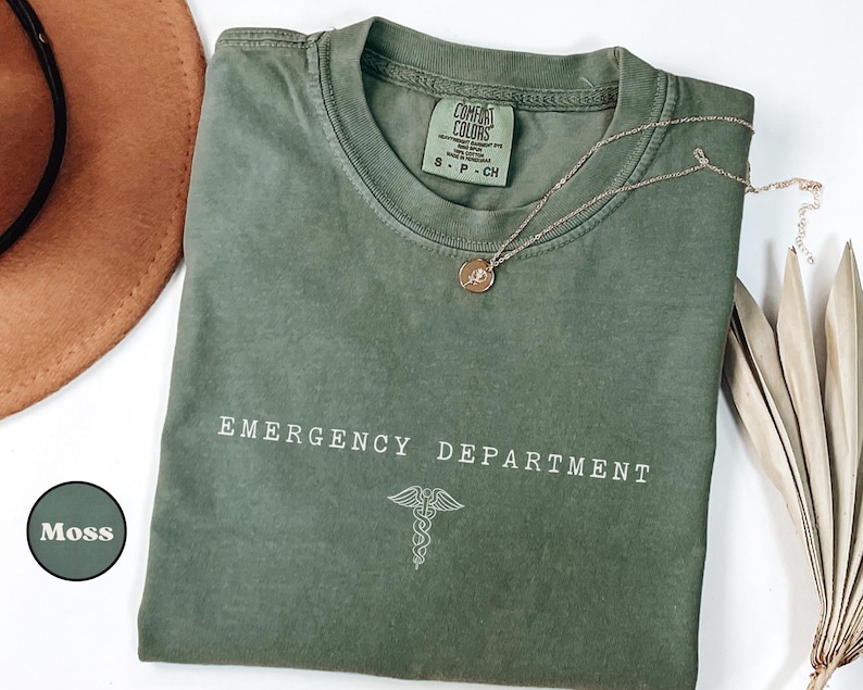 May include: A moss green crewneck t-shirt with the text EMERGENCY DEPARTMENT printed in white letters above a white caduceus symbol. The shirt features a Comfort Colors brand label at the inner neckline and a small circular color swatch labeled Moss.