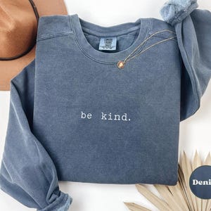 May include: A dark blue sweatshirt with the text "be kind." embroidered on the front. The sweatshirt has a distressed look and is made of a soft, comfortable fabric.
