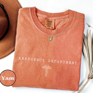 May include: A terracotta-colored t-shirt with a crew neckline and a Comfort Colors label. The shirt features the white text EMERGENCY DEPARTMENT and a caduceus symbol printed on the front. A small circular inset shows the color name Yam.