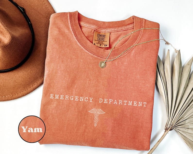 May include: A terracotta-colored t-shirt with a crew neckline and a Comfort Colors label. The shirt features the white text EMERGENCY DEPARTMENT and a caduceus symbol printed on the front. A small circular inset shows the color name Yam.