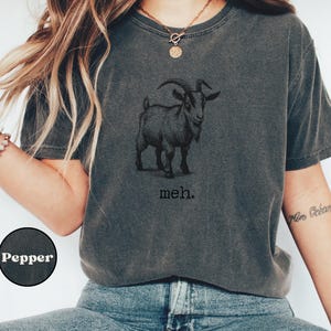May include: A dark gray t-shirt with a black and white illustration of a goat and the text "meh."