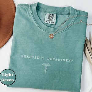 May include: A light green short-sleeve t-shirt with a crew neckline. The chest features the white text EMERGENCY DEPARTMENT above a white caduceus symbol. The interior neck label reads COMFORT COLORS, 100% COTTON, MADE IN HONDURAS, S P CH.