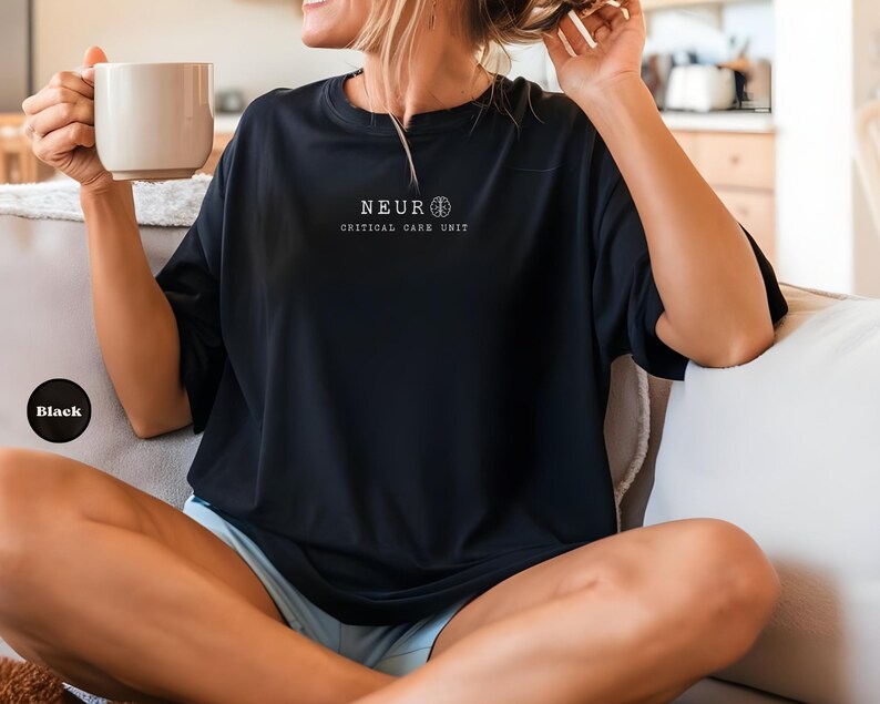May include: Black t-shirt with the text "NEUR CRITICAL CARE UNIT" in white. The shirt has a crew neck and short sleeves. A black circle with the word "Black" is in the lower left corner. The person is holding a beige mug.