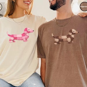 May include: Ivory and espresso-colored t-shirts featuring a dachshund design. The ivory shirt has a pink patchwork dog, while the espresso shirt has a brown and tan patchwork dog. The shirts are casual and comfortable.
