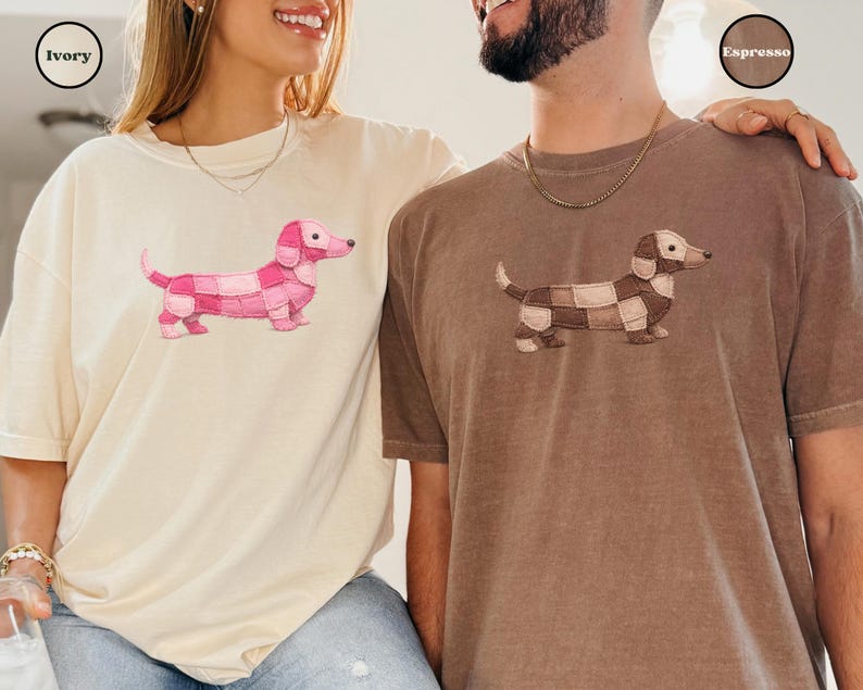 May include: Ivory and espresso-colored t-shirts featuring a dachshund design. The ivory shirt has a pink patchwork dog, while the espresso shirt has a brown and tan patchwork dog. The shirts are casual and comfortable.