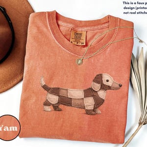 May include: A coral-colored t-shirt featuring a dachshund dog design made of faux patchwork. The shirt has a crew neck and short sleeves. A gold necklace with a pendant is draped on the shirt. The text "This is a faux patchwork design (printed effect), not real stitched fabric." is visible.