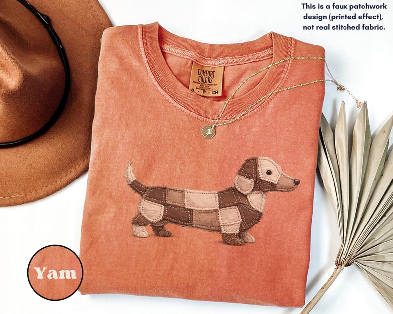 May include: A coral-colored t-shirt featuring a dachshund dog design made of faux patchwork. The shirt has a crew neck and short sleeves. A gold necklace with a pendant is draped on the shirt. The text "This is a faux patchwork design (printed effect), not real stitched fabric." is visible.