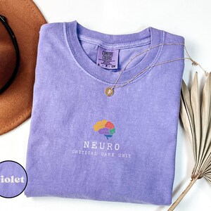 May include: A violet Comfort Colors t-shirt with a crew neck and short sleeves. The shirt features a colorful brain graphic and the text "NEURO CRITICAL CARE UNIT". A gold necklace and a brown hat are also in the image.
