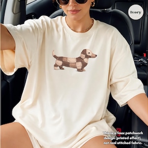 May include: Ivory-colored t-shirt featuring a dachshund dog illustration with a patchwork design. The dog is depicted in shades of brown and tan. The text on the shirt reads: "This is a faux patchwork design (printed effect), not real stitched fabric."