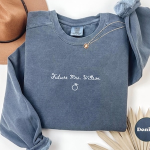 May include: A denim blue sweatshirt with the embroidered text "Future Mrs. Willson" and a ring graphic. The long-sleeved crew neck shirt has a relaxed fit and is made of a soft material. A gold necklace is draped across the shirt.