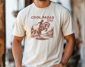 May include: Cream-colored t-shirt featuring a brown graphic of a cowboy riding a bucking horse, with the text "COOL PAPAS Club" in brown and red. The design includes a ranch scene with trees and a fence.