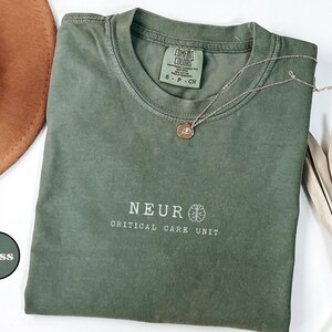 May include: A moss green t-shirt with the text "NEUR CRITICAL CARE UNIT" in white. A gold necklace with a pendant rests on the shirt. The shirt is folded and placed on a white surface, with a brown hat and dried leaves in the background.