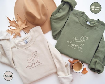 Puede incluir: Sudaderas de color arena y verde militar con un diseño de perro salchicha y el nombre "Charlie". Las sudaderas se muestran con un sombrero marrón, una hoja de otoño y una taza de café. También está presente el texto "Life's better with a dachshund".
