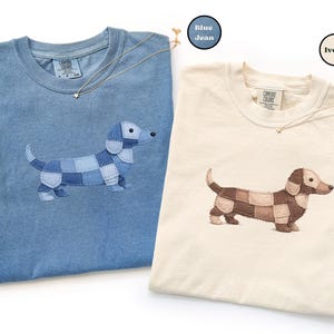 May include: Two t-shirts, one blue and one ivory, each featuring an embroidered dachshund dog design. The dog's body is made of patchwork squares. Each shirt has a Comfort Colors tag. A gold necklace with a star pendant is draped on each shirt.