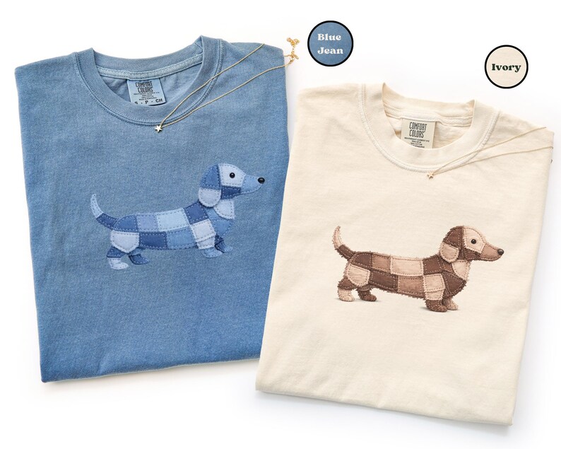 May include: Two t-shirts, one blue and one ivory, each featuring an embroidered dachshund dog design. The dog's body is made of patchwork squares. Each shirt has a Comfort Colors tag. A gold necklace with a star pendant is draped on each shirt.