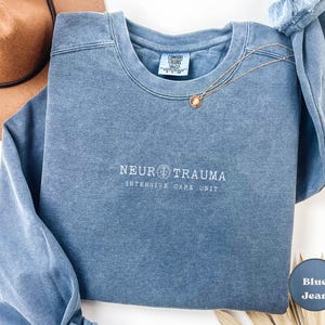 May include: A blue jean-colored sweatshirt with the words "NEURO TRAUMA INTENSIVE CARE UNIT" in white lettering. A gold necklace with a circular pendant is draped across the front. A brown hat is visible in the upper left corner.
