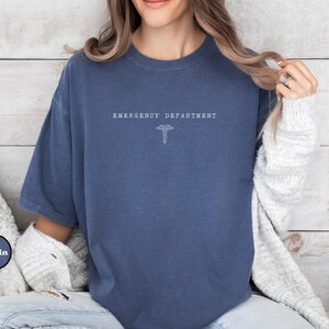 May include: A navy blue short-sleeve t-shirt featuring the white text EMERGENCY DEPARTMENT centered on the chest above a small white caduceus symbol. The shirt has a crew neckline and a relaxed fit, with a visible Demin logo in the corner.