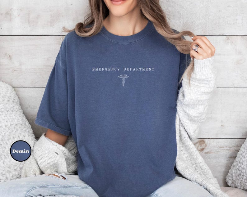 May include: A navy blue short-sleeve t-shirt featuring the white text EMERGENCY DEPARTMENT centered on the chest above a small white caduceus symbol. The shirt has a crew neckline and a relaxed fit, with a visible Demin logo in the corner.