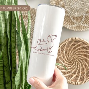 May include: A white 20 oz. skinny tumbler featuring a brown outline of a dachshund dog with the name "Charlie" and the phrase "Life's Better with a Dachshund." The tumbler is held in a person's hand.