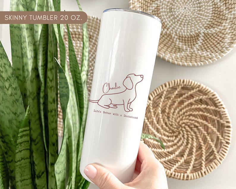 May include: A white 20 oz. skinny tumbler featuring a brown outline of a dachshund dog with the name "Charlie" and the phrase "Life's Better with a Dachshund." The tumbler is held in a person's hand.
