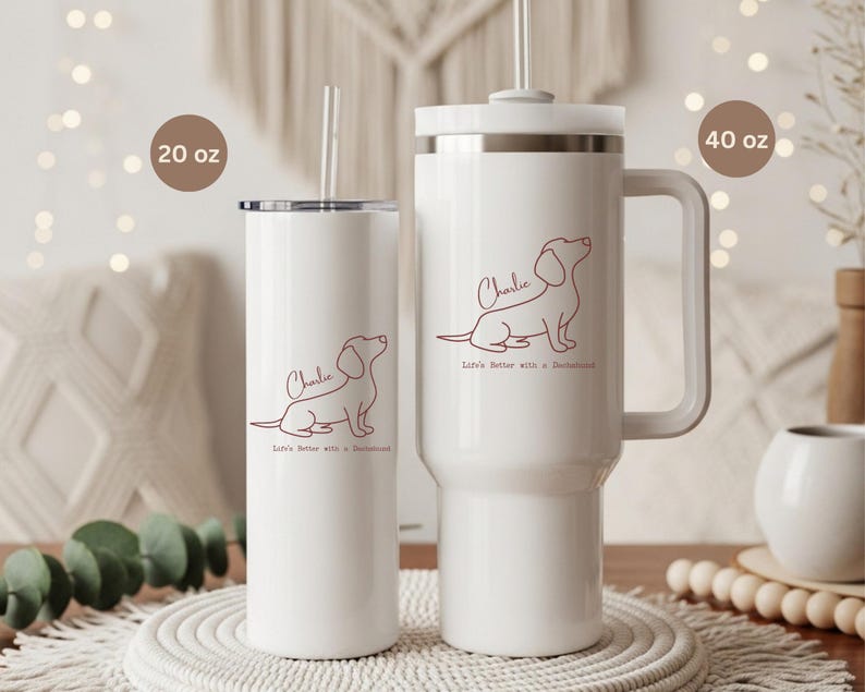 May include: Two white insulated tumblers, one 20 oz and one 40 oz, with a dachshund outline and the name "Charlie" in a script font. The text "Life's better with a dachshund" is below the dog illustration. Both tumblers have a straw.