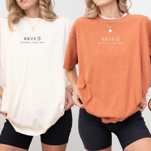 May include: Two oversized t-shirts, one ivory and one yam-colored, each featuring the text "NEUR CRITICAL CARE UNIT" in black. The shirts are paired with black shorts. The ivory shirt is labeled "Ivory" and the yam shirt is labeled "Yam".