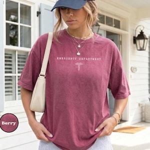 May include: A berry-colored, short-sleeved t-shirt featuring the white text "EMERGENCY DEPARTMENT" and a medical caduceus symbol centered on the chest. The shirt has a relaxed fit and is worn with a light-colored shoulder bag and a baseball cap.