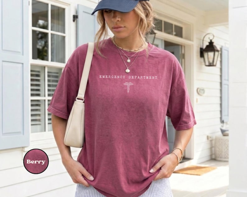 May include: A berry-colored, short-sleeved t-shirt featuring the white text "EMERGENCY DEPARTMENT" and a medical caduceus symbol centered on the chest. The shirt has a relaxed fit and is worn with a light-colored shoulder bag and a baseball cap.