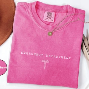 May include: A bright pink short-sleeved t-shirt with a crew neckline. The chest features the white text EMERGENCY DEPARTMENT above a white caduceus symbol. The inner neck label reads Comfort Colors, 100% cotton, size S, made in Honduras.
