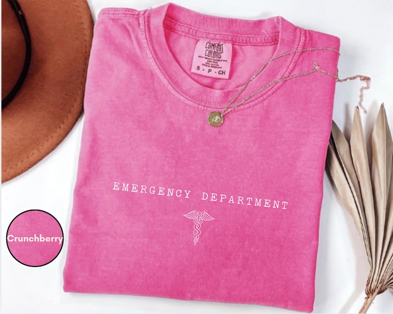 May include: A bright pink short-sleeved t-shirt with a crew neckline. The chest features the white text EMERGENCY DEPARTMENT above a white caduceus symbol. The inner neck label reads Comfort Colors, 100% cotton, size S, made in Honduras.