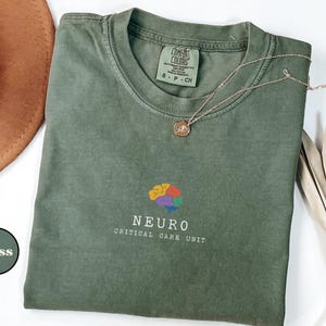 May include: A moss green t-shirt with a crew neck, featuring a colorful brain graphic and the text "NEURO CRITICAL CARE UNIT." A gold necklace rests on the shirt. A brown hat and dried plant are in the background.