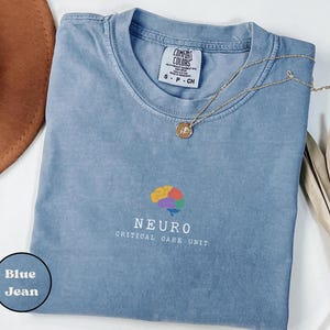 May include: A blue jean Comfort Colors t-shirt with the word "NEURO" and a colorful brain graphic. The words "CRITICAL CARE UNIT" are below the word "NEURO". A gold necklace with a pendant is on the shirt.