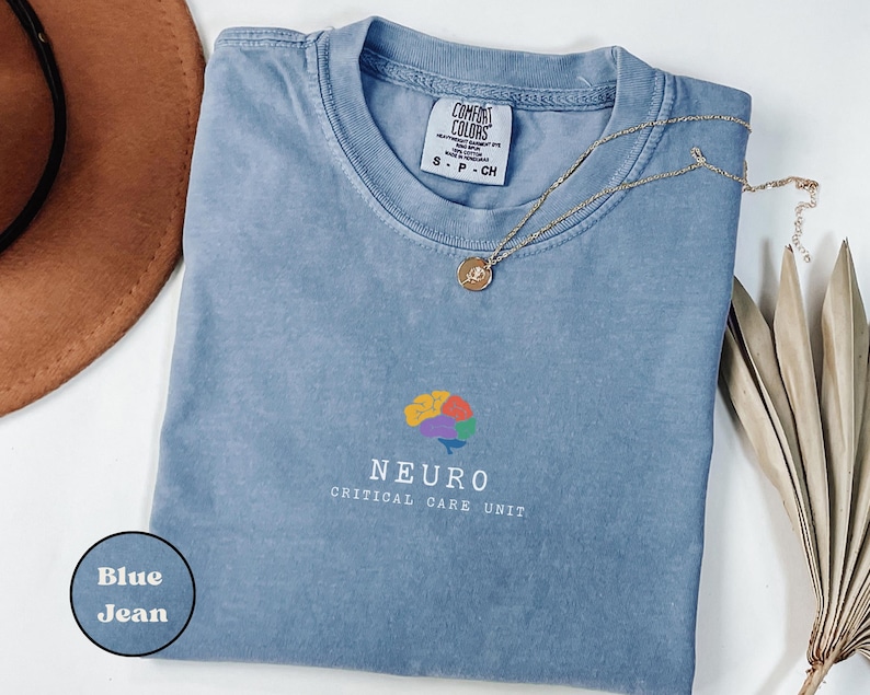 May include: A blue jean Comfort Colors t-shirt with the word "NEURO" and a colorful brain graphic. The words "CRITICAL CARE UNIT" are below the word "NEURO". A gold necklace with a pendant is on the shirt.