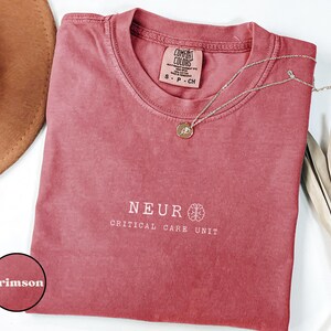 May include: Crimson-colored t-shirt with the words "NEUR CRITICAL CARE UNIT" printed in white. The shirt has a crew neck and short sleeves. A gold necklace with a pendant rests on the shirt. A brown hat and dried leaves are in the background.