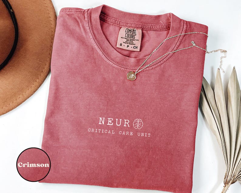 May include: Crimson-colored t-shirt with the words "NEUR CRITICAL CARE UNIT" printed in white. The shirt has a crew neck and short sleeves. A gold necklace with a pendant rests on the shirt. A brown hat and dried leaves are in the background.